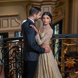 Elegant wedding couple portrait at a luxury venue by The Glorious Memories A groom in a black suit and a bride in a gold embellished gown pose together at an ornate indoor wedding venue railing.