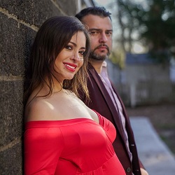 The Glorious Memories | Baby Shower Photography in New Hyde Park, NY A smiling woman in a red off-shoulder dress posing outdoors beside a man in a suit, with a softly blurred background.